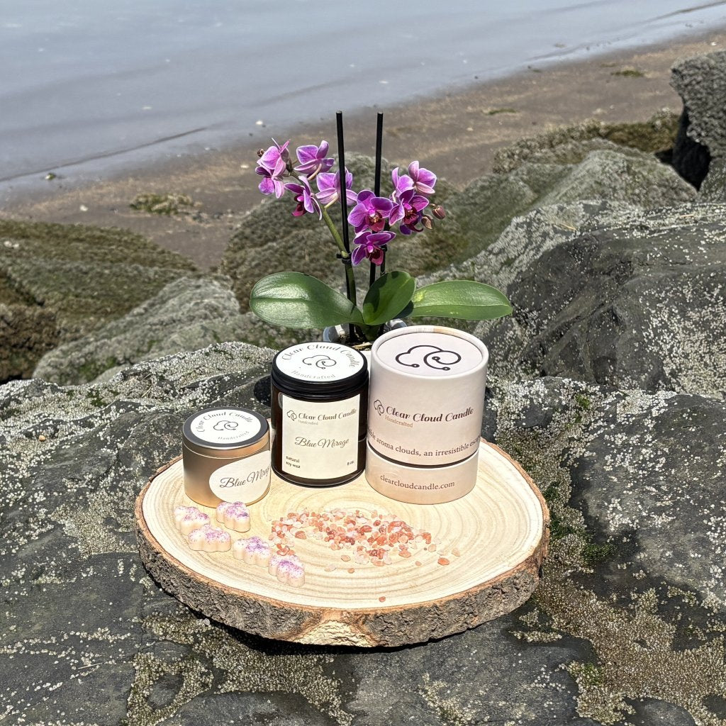 amber glass candle jar. golden tin candle container. paper packaging tube with company name and logo. cloud-shaped wax melts. orchid vase in background. on rocky waterfront