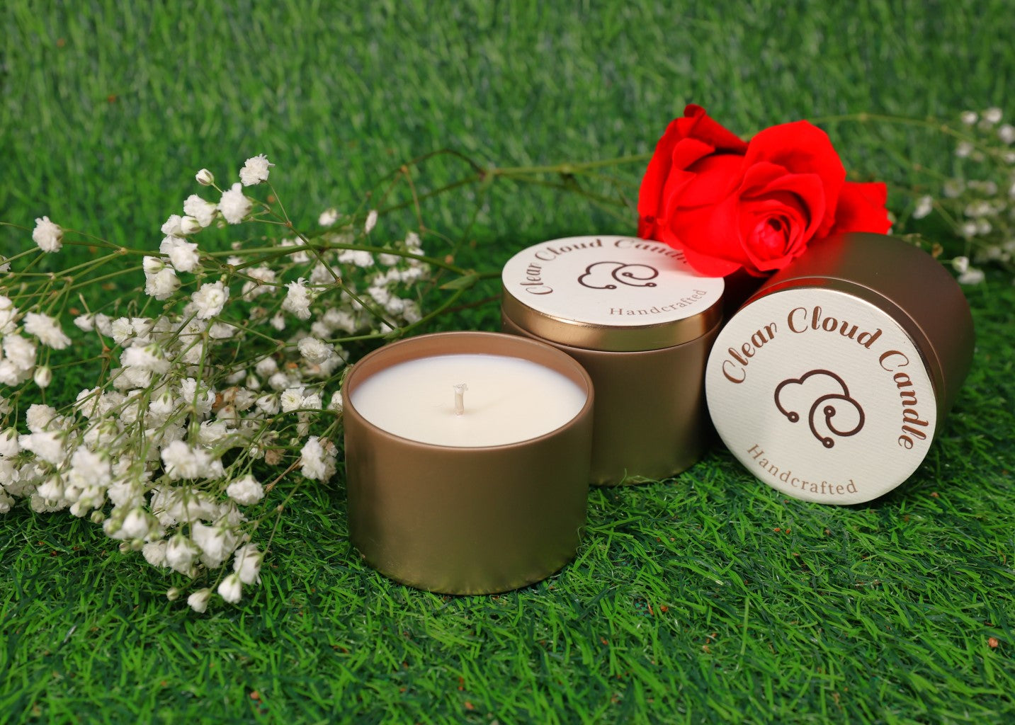 rose gold tin container with white candle. 2 rose gold tin candle containers with tops. clear cloud candle logo on the tops. on green grass background with baby breaths flowers and a red rose