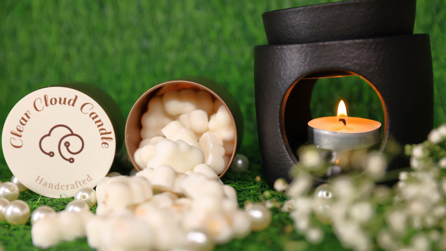 white cloud-shaped wax melts spilling out from rose gold tin container. container cover with clear cloud candle logo. tealight candle lit under a black wax Melter. green grass background
