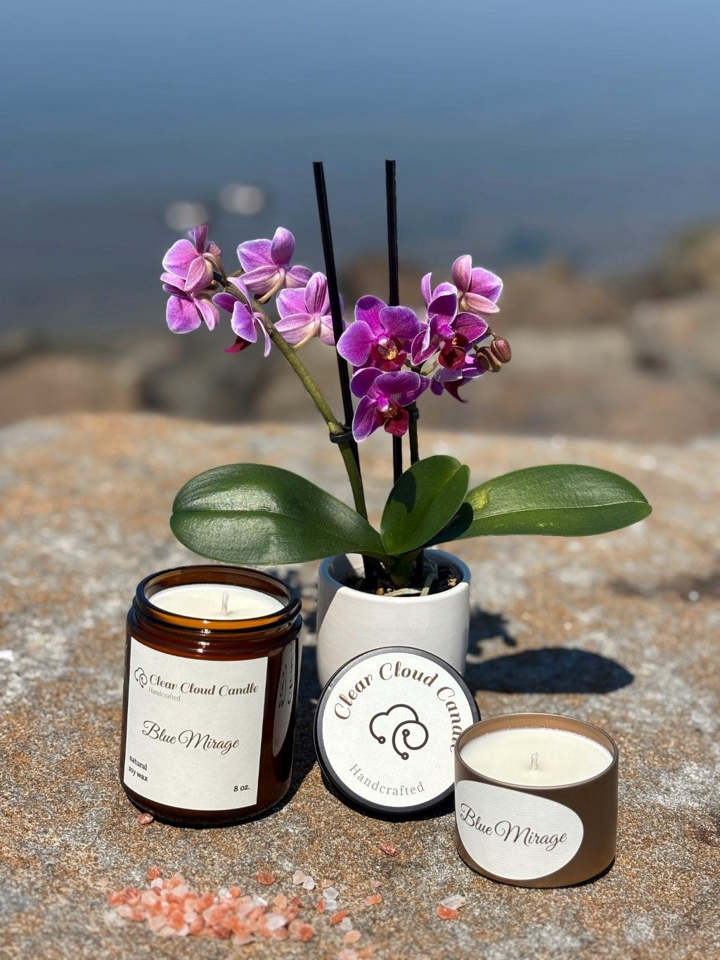 candle in amber glass jar with black cover. clear cloud candle round logo on the cover. label reads Blue Mirage. candle in golden rose tin container. label reads "blue mirage". fresh orchid plant and flower behind. on rocky ground with blurred ocean background.