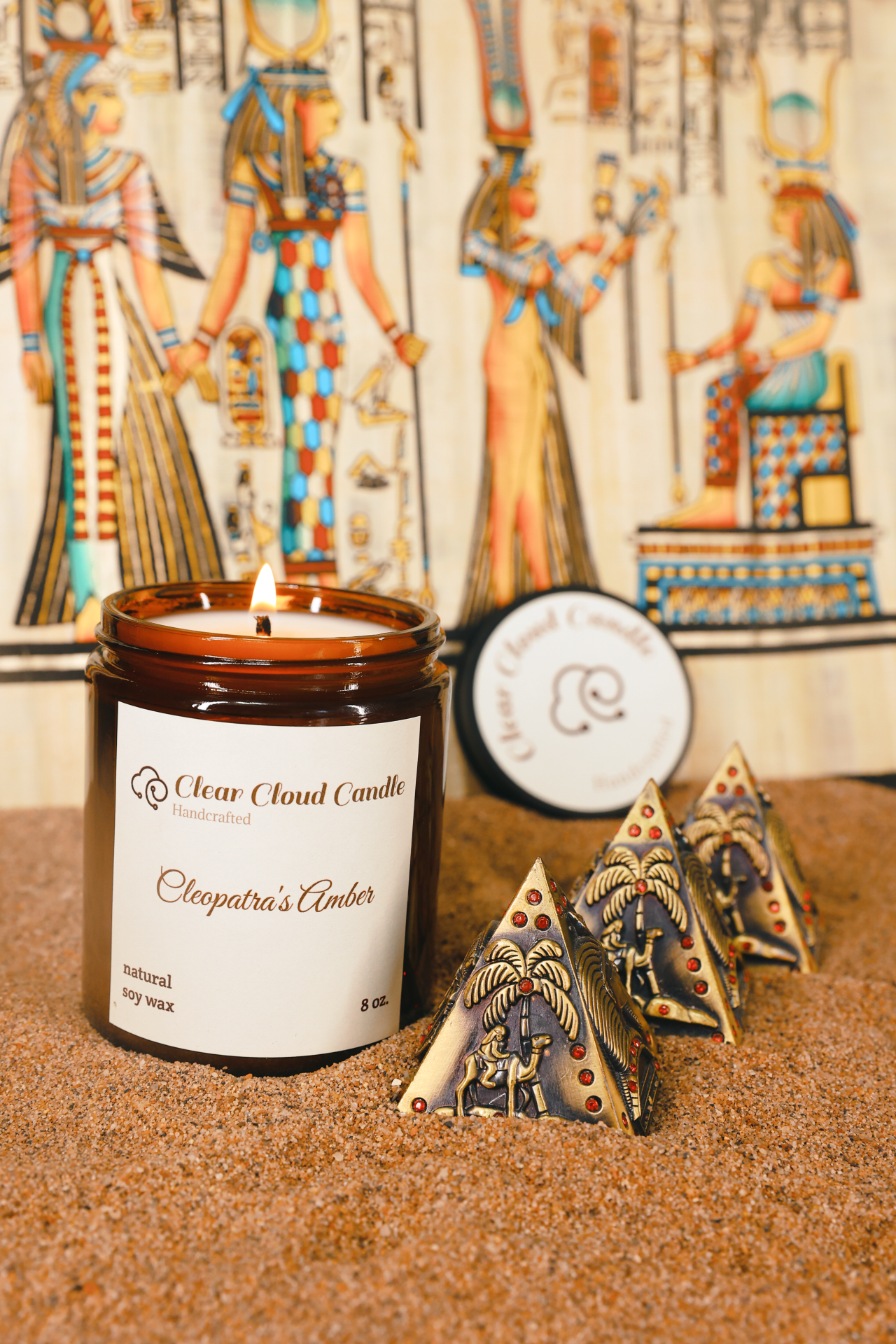 lit scented candle in amber glass jar, scent name: cleopatra's amber. ancient Egyptian props, mini pyramids, sand ground
