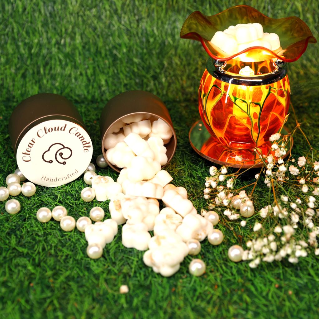 white cloud-shaped wax melts spilling out from rose gold tin container. side-laying tin container with clear cloud candle logo on the cover. amber color glass light-bulb-operated wax Melter. green grass background with white flowers.