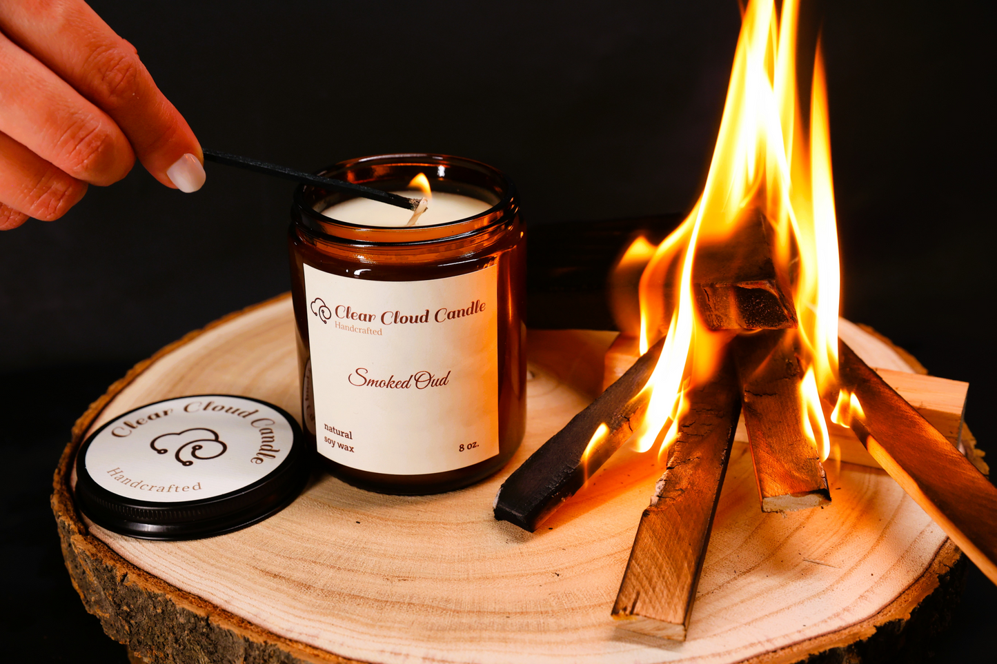 hand lighting candle in amber glass jar, light cream label. wood on fire. smoke. flames. cent name: smoked oud