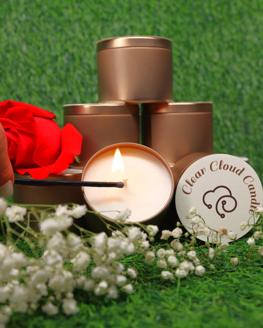 partial hand lighting a white candle. the staked golden rose containers. on container with clear cloud candle logo on the top, green grass background with white flowers and a red rose.