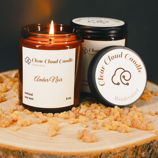 A soy wax candle with the label 'Amber Noir' burning on a wooden surface, surrounded by raw amber pieces and the lid of the jar with the brand 'Clean Cloud Candle' visible in the background.