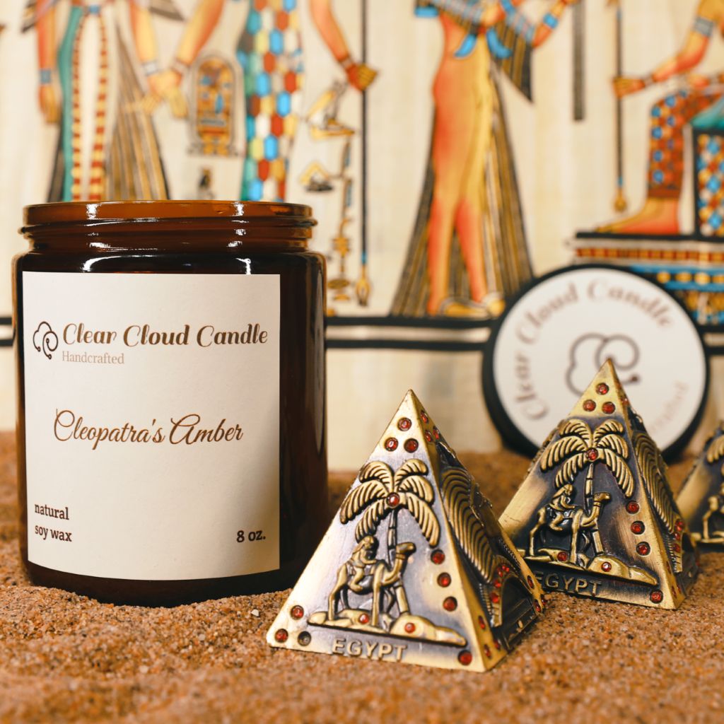 Clear Cloud Candle jar with 'Cleopatra's Amber' scent, accompanied by decorative Egyptian-themed pyramids on a sandy surface.