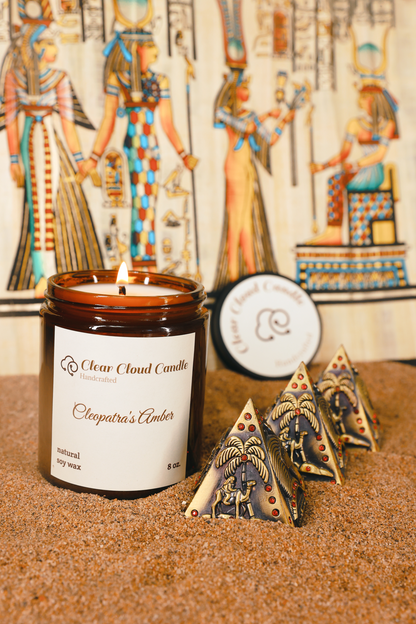 lit scented candle in amber glass jar, scent name: cleopatra's amber. ancient Egyptian props, mini pyramids, sand ground