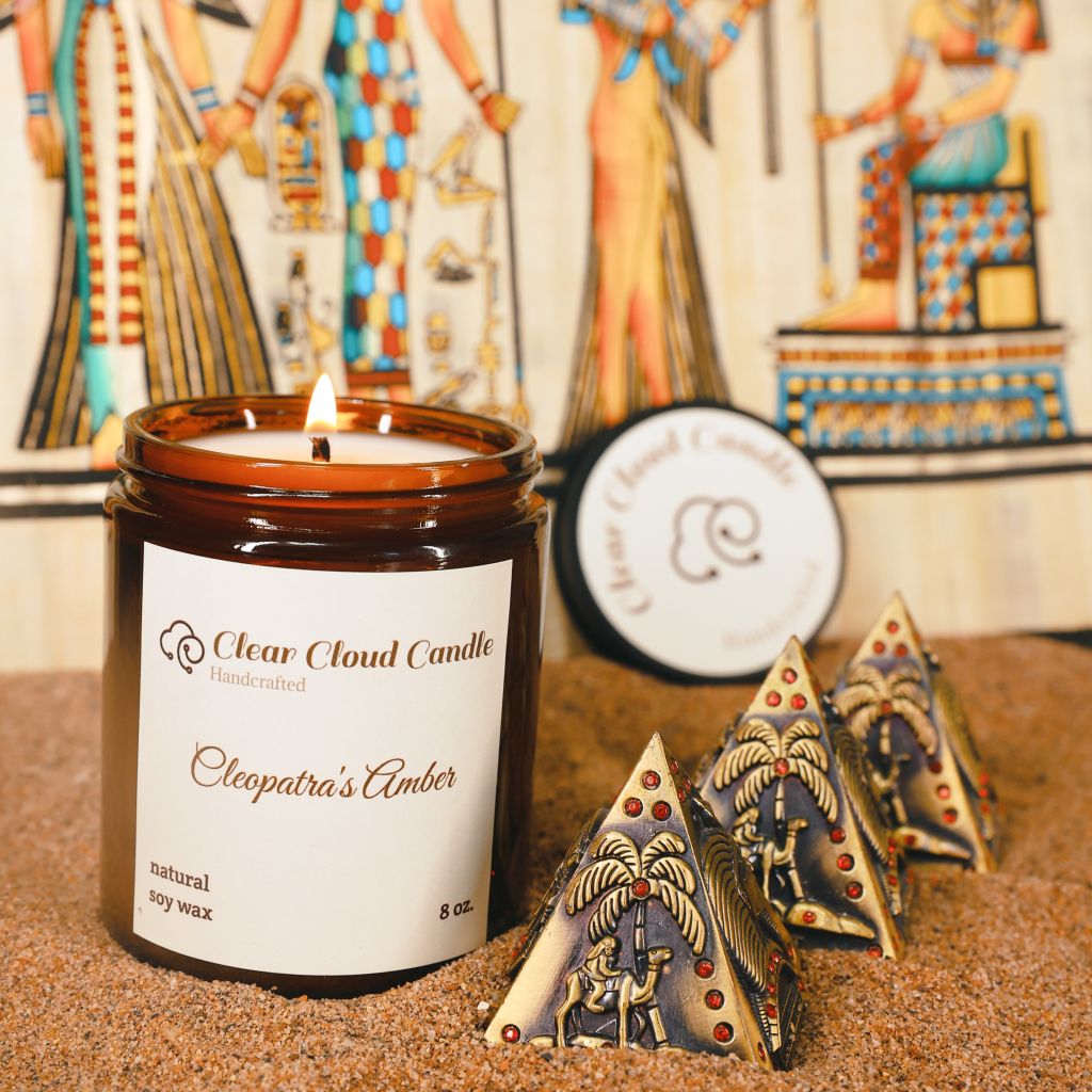 Cleopatra's Amber scented candle by 'Clear Cloud Candle' Co., with decorative pyramids on sand surface.