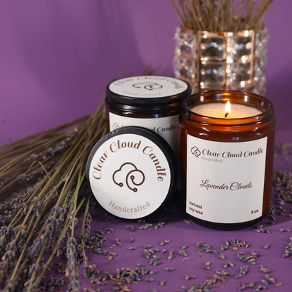 Two candles from Clear Cloud Candle Co., one lit with a visible flame, surrounded by dried lavender and a glass vase in the background.