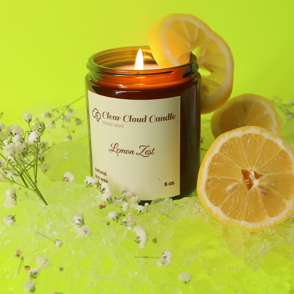 Lemon Zest scented candle by "Clear Cloud Candle" Co., lit and surrounded by lemon slices, lemon peel, and small white flowers on ice with yellow background.