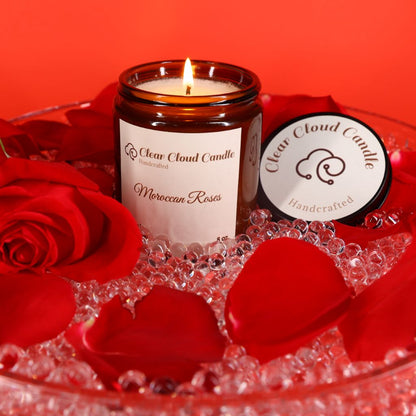A lit Moroccan Roses scented candle in amber jar, with "clear cloud candle" Co. on the label, surrounded by red rose petals and crystals on a red background.