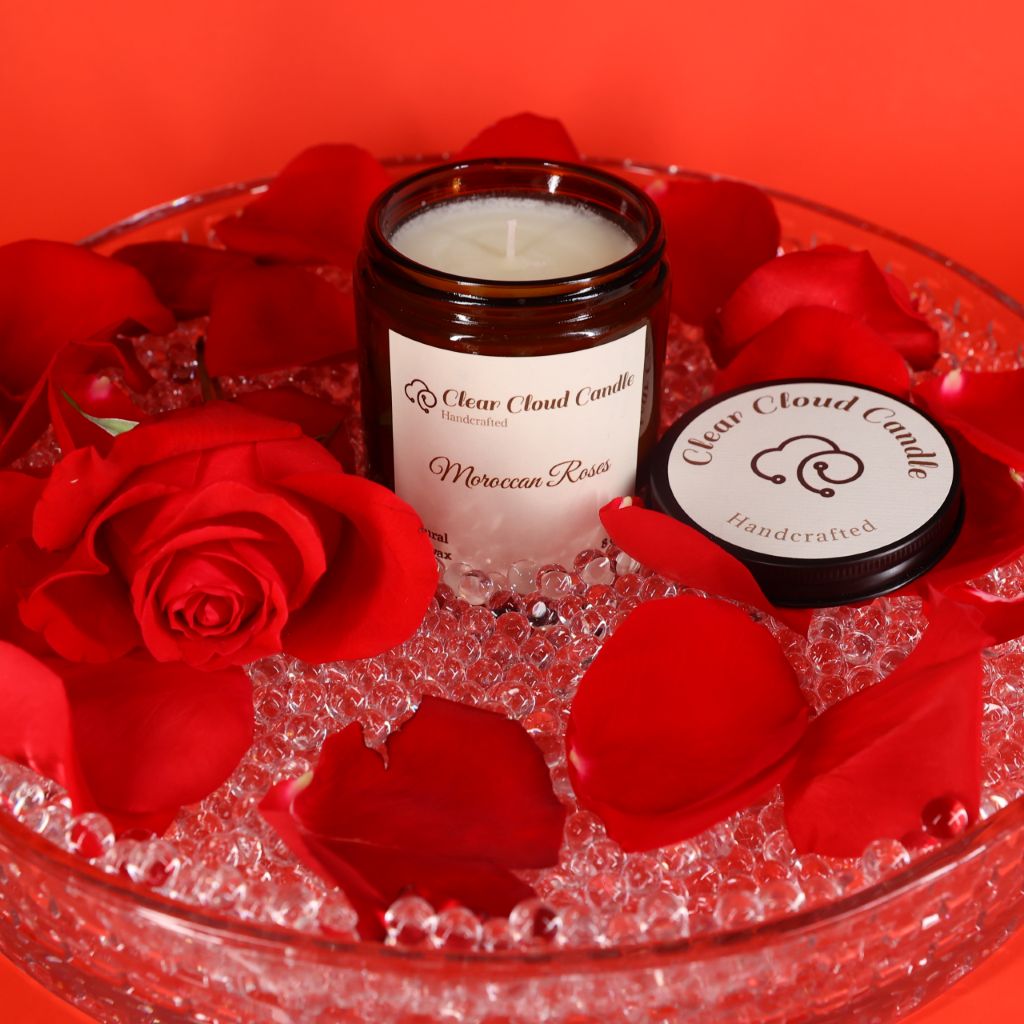 A lit Moroccan Roses scented candle in amber jar, with "clear cloud candle" Co. on the label, surrounded by red rose petals and crystals on a red background.