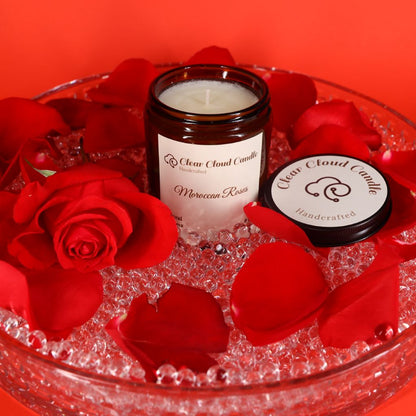 A lit Moroccan Roses scented candle in amber jar, with "clear cloud candle" Co. on the label, surrounded by red rose petals and crystals on a red background.