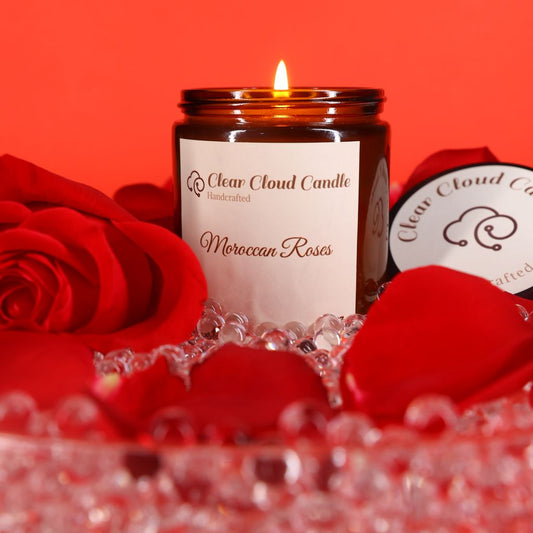A lit Moroccan Roses scented candle in amber jar, with "clear cloud candle" Co. on the label, surrounded by red rose petals and crystals on a red background.