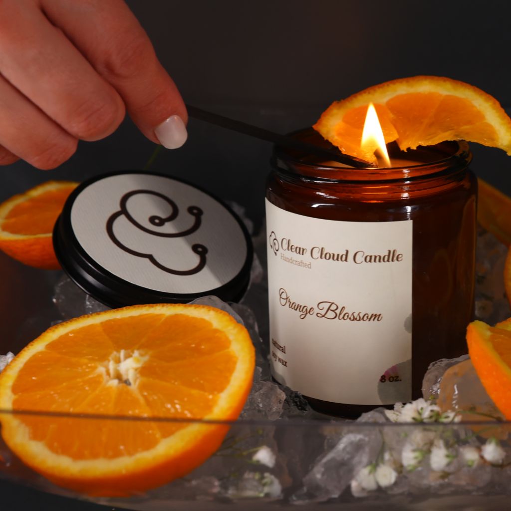 Amber color Candle jar with cream labeled reads 'Orange Blossom' scent from "Clear Cloud Candle" Co. with a hand lighting the candle. surrounded by sliced oranges.