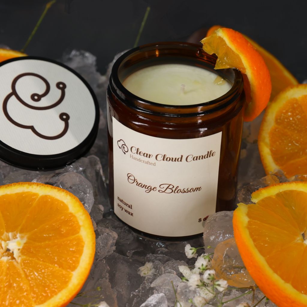 amber color candle Jar with cream label from 'Clear Cloud Candle' Co. Orange Blossom scent with sliced oranges and ice cubes on a dark surface