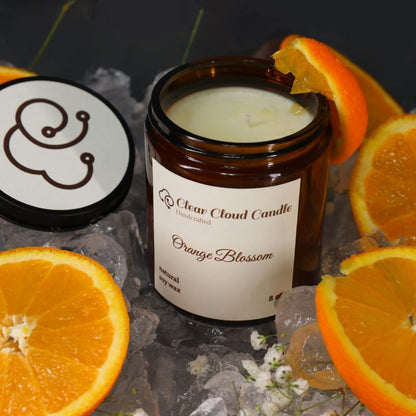 amber color candle Jar with cream label from 'Clear Cloud Candle' Co. Orange Blossom scent with sliced oranges and ice cubes on a dark surface