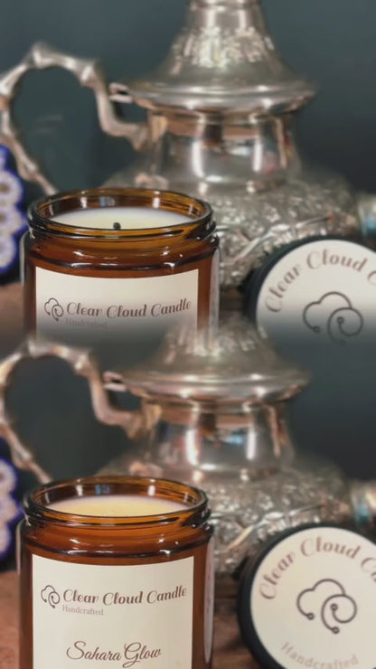 mirrored view of candle being lit. candle in amber glass jar with cream label. Label reads the scent name "Sahara Glow". clear cloud candle logo on the label.