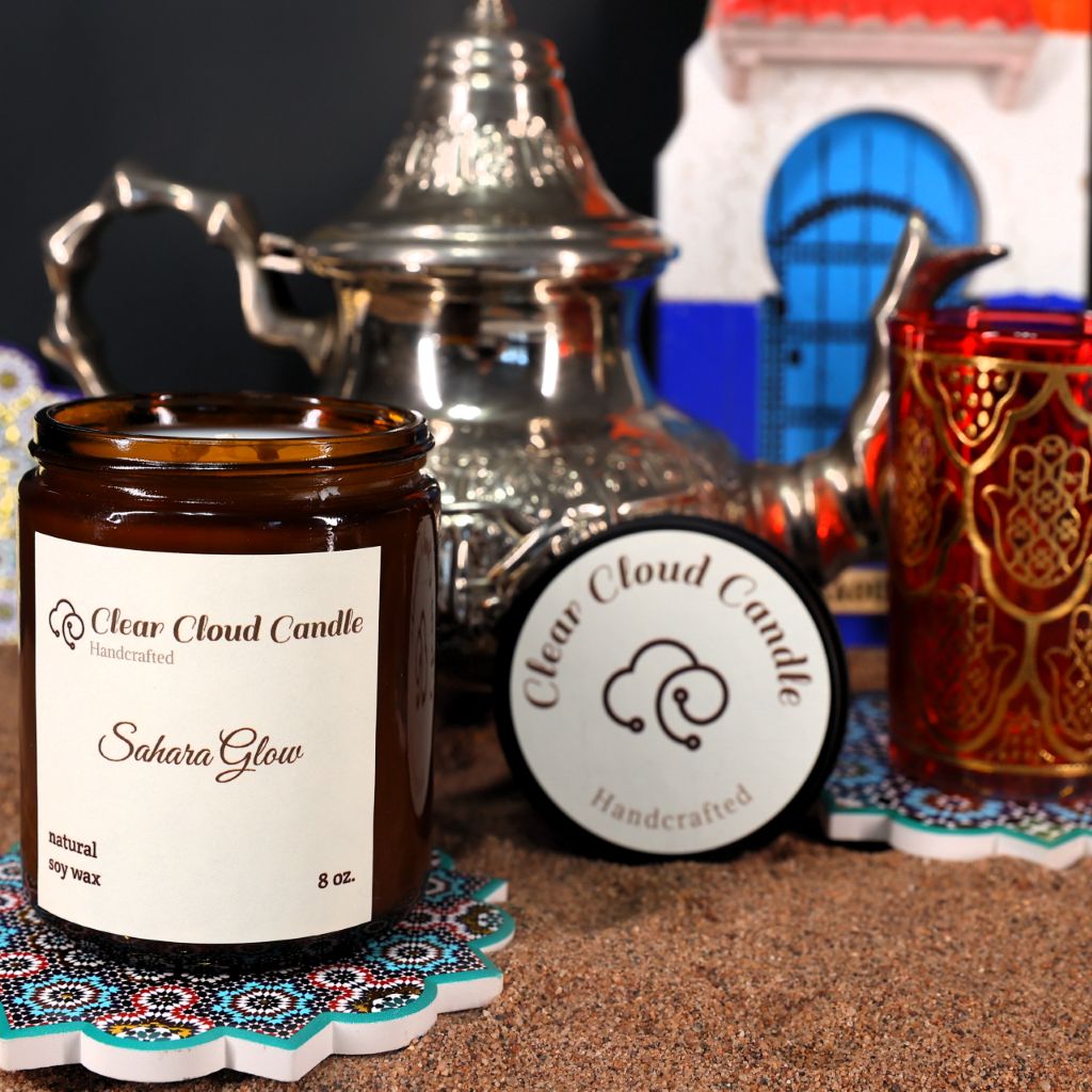 Clear Cloud Candle Co. in amber jar with scent name "Sahara Glow" with Moroccan decorations, teapot, tea cups and on a sandy background