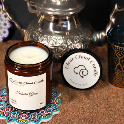 Scented candle with scent name "Sahara Glow" from "Clear Cloud Candle" Co. in amber jar with lid and decorative coaster on sand background