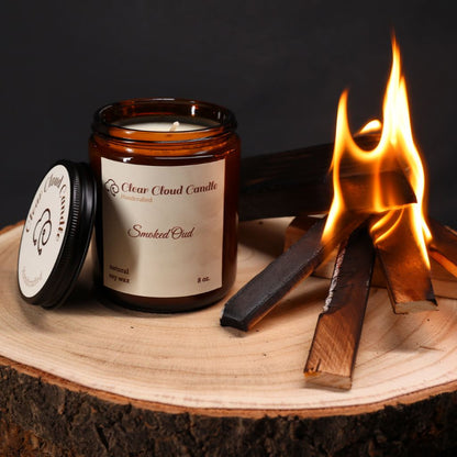 Smoked Oud scented candle in amber glass jar with cream color label from "Clear Cloud Candle" Co. on a wooden surface next to burning wood.