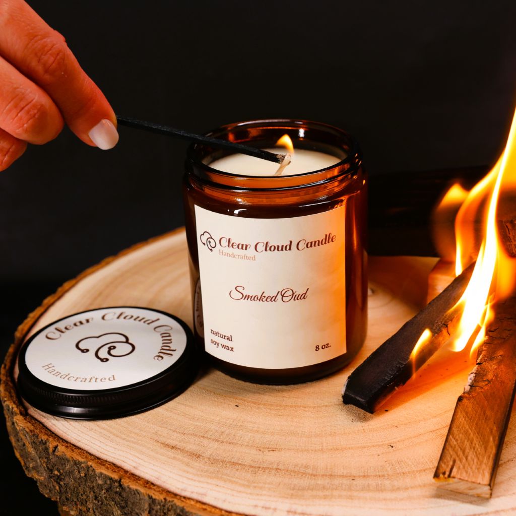 amber color jar with "Clear Cloud Candle" Co. label, scent name is "Smoked Oud" being lit on a wooden surface with firewood and a dark background