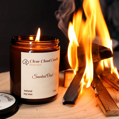 lit candle in amber glass jar, light cream label. wood on fire. smoke. flames. cent name: smoked oud. By "Clear Cloud Candle" Co.