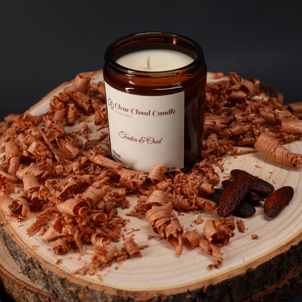 Candle in amber color jar with "Clear Cloud Candle" Co. Label on a wooden surface with wood shavings and tonka nuts on black background.