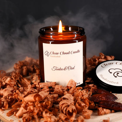 A lit candle labeled 'Tonka & Oud' by Clear Cloud Candle, surrounded by wooden shavings, with smoke rising in the air.