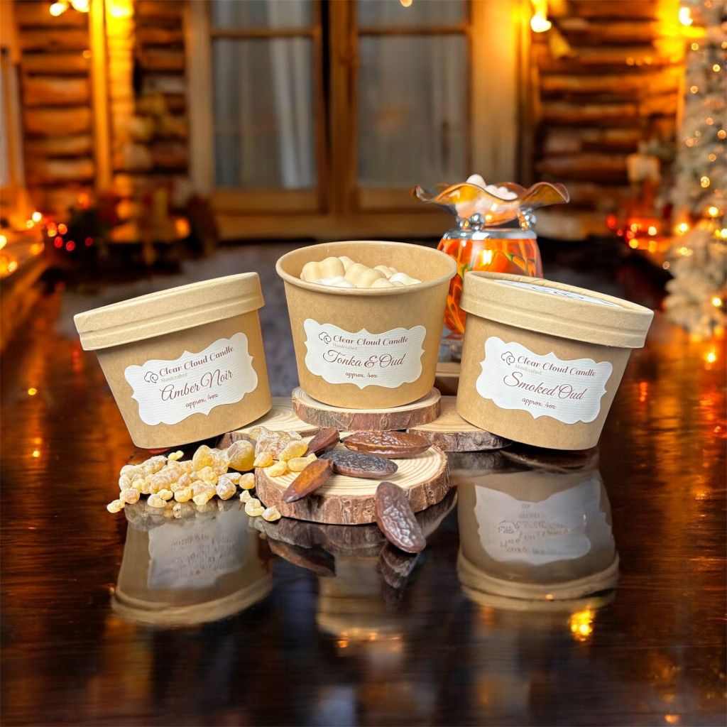 3 kraft-paper colored containers from "Clear Cloud Candle" with lids and labels reading various scents names on a wooden surface, surrounded by tonka beans, amber pieces and a wax Melter in the background. one of the containers is uncovered with cloud-shaped wax melts visible inside.
